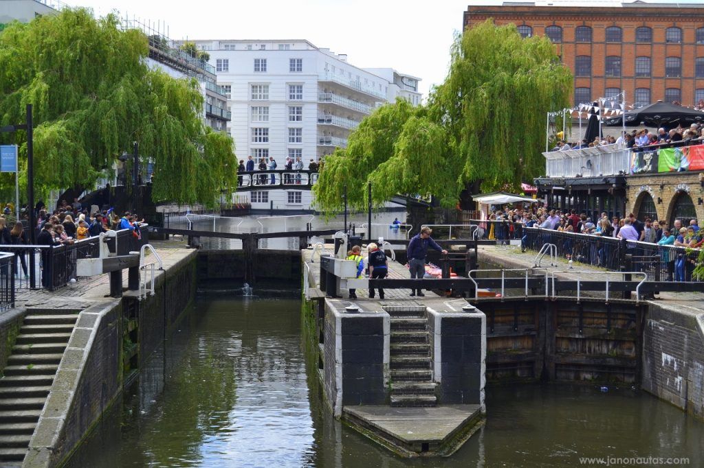 El canal de Camden Vista del canal junto a Camden Market en Londres, con esclusas de agua y un puente peatonal, rodeado de vegetación y gente paseando.